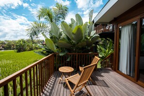 Una terraza de madera con una mesa y sillas, rodeada de abundante vegetación y vistas a un campo de arroz.