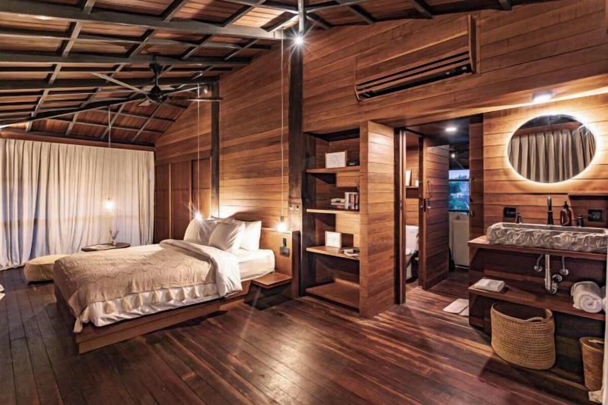 A wooden-themed bedroom with a bed, bedside tables, a built-in wardrobe, and a bathroom with a round mirror and sink.