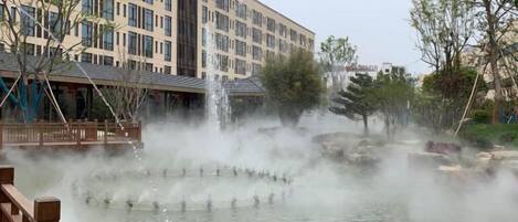 A water feature with multiple fountains, a wooden deck, and a building with a modern facade in the background.