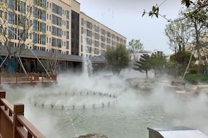 A water feature with multiple fountains, a wooden deck, and a building with a modern facade in the background. - Heye Hotel (Anji)