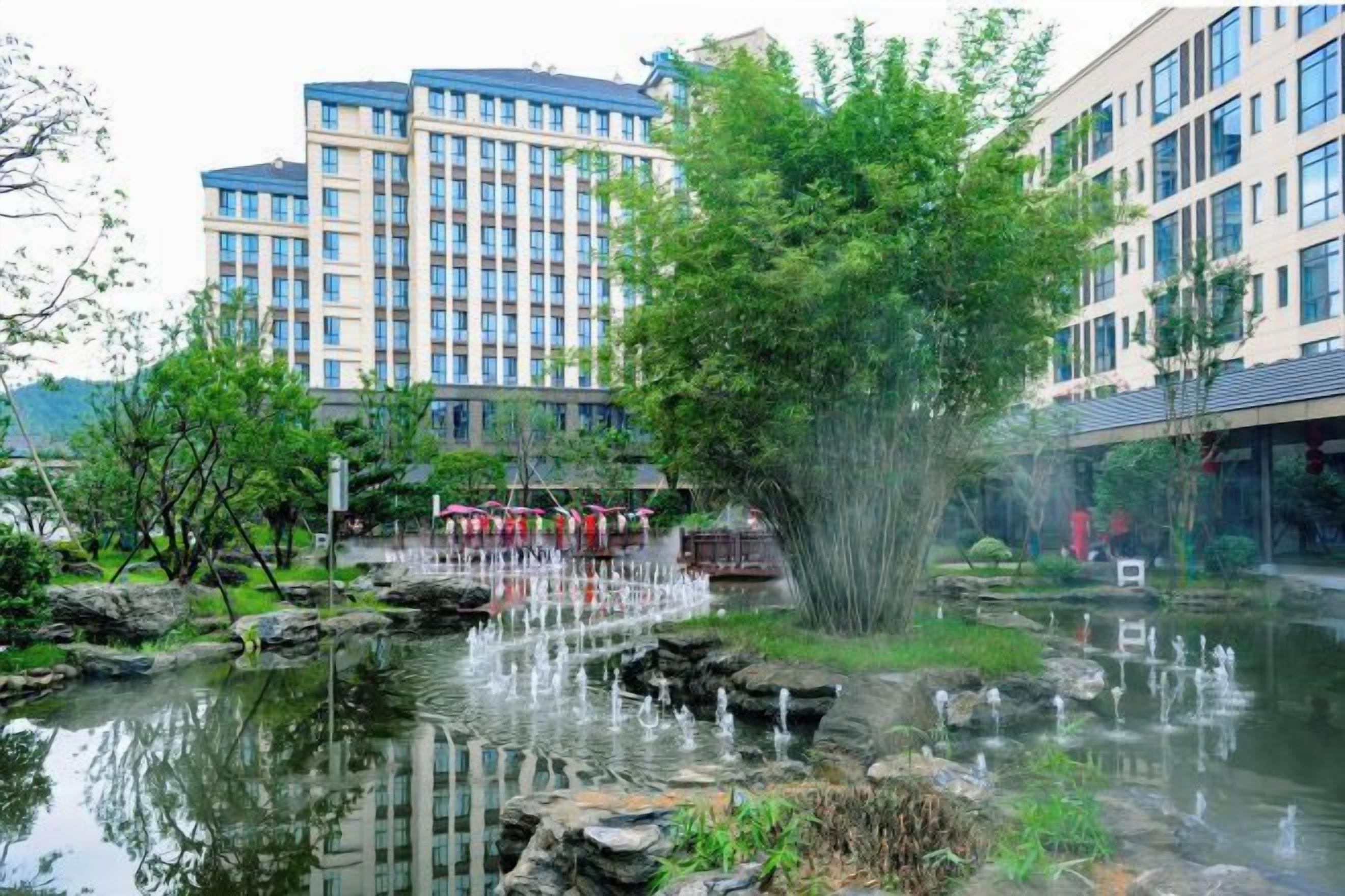 A water feature with fountains, surrounded by greenery and buildings in the background.