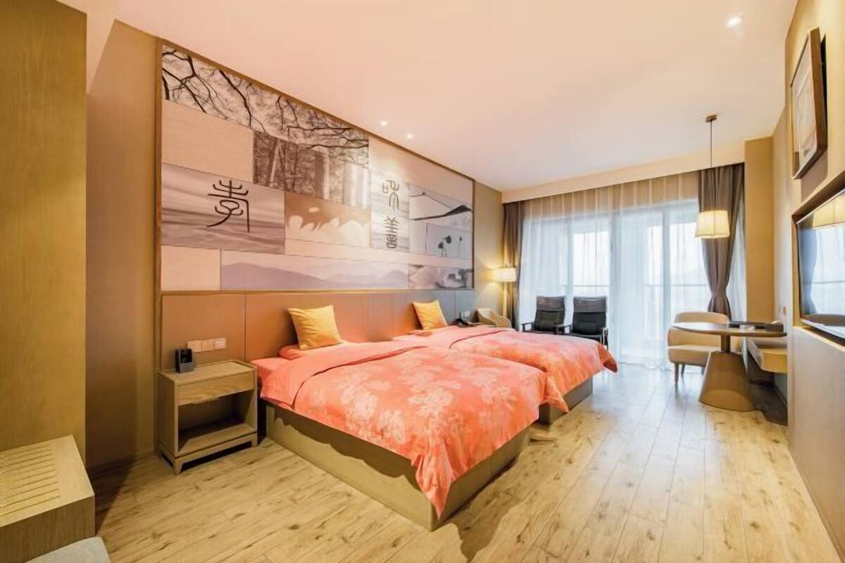 A hotel room with a large bed, a desk, and a chair. There is a wall with artwork and a window with curtains.
