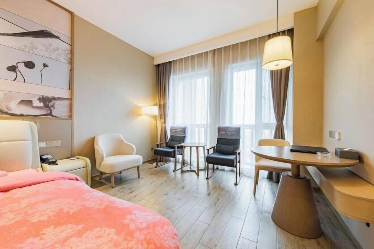 A hotel room with a bed, a desk, two chairs, a small table, a lamp, and a window with curtains.