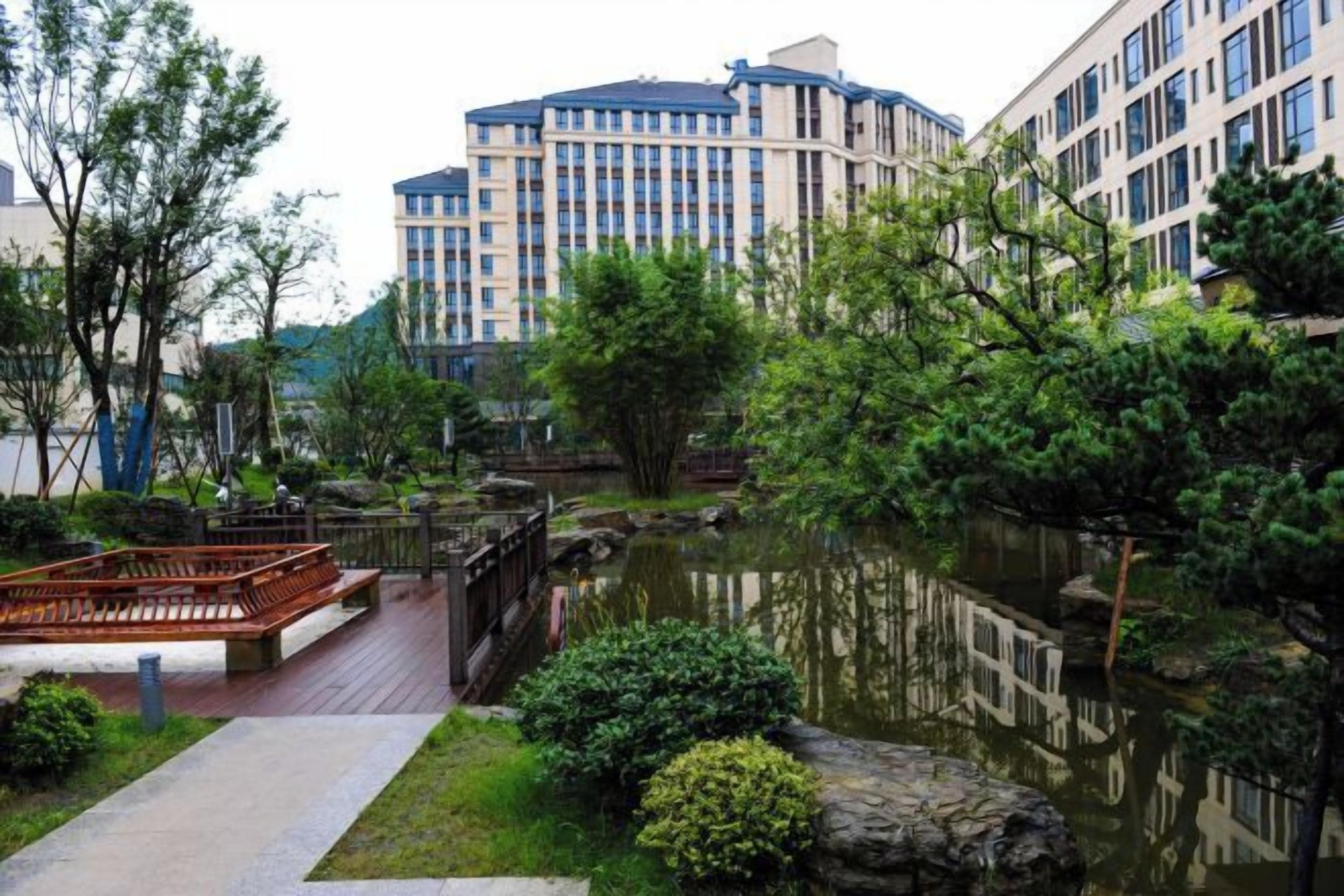 A landscaped area with a pond, wooden bridge, and buildings in the background.