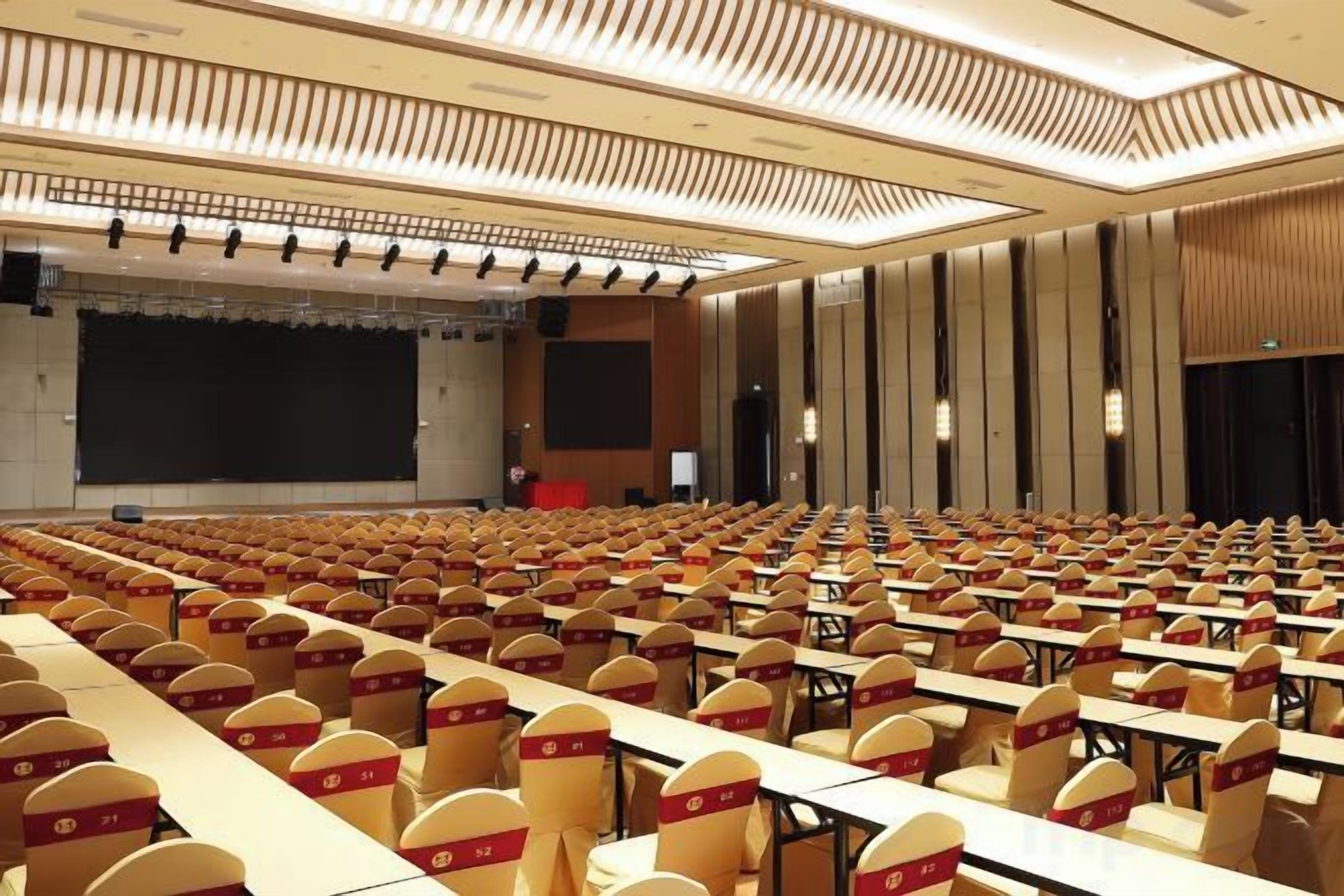 Een congreszaal met rijen tafels en stoelen, een podium en een groot scherm.