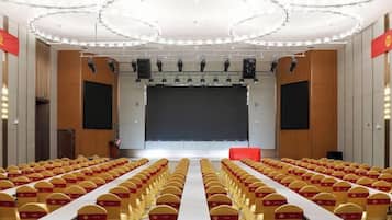 Un salón de conferencias con filas de mesas y sillas, un escenario con una pantalla y un techo alto de color blanco.