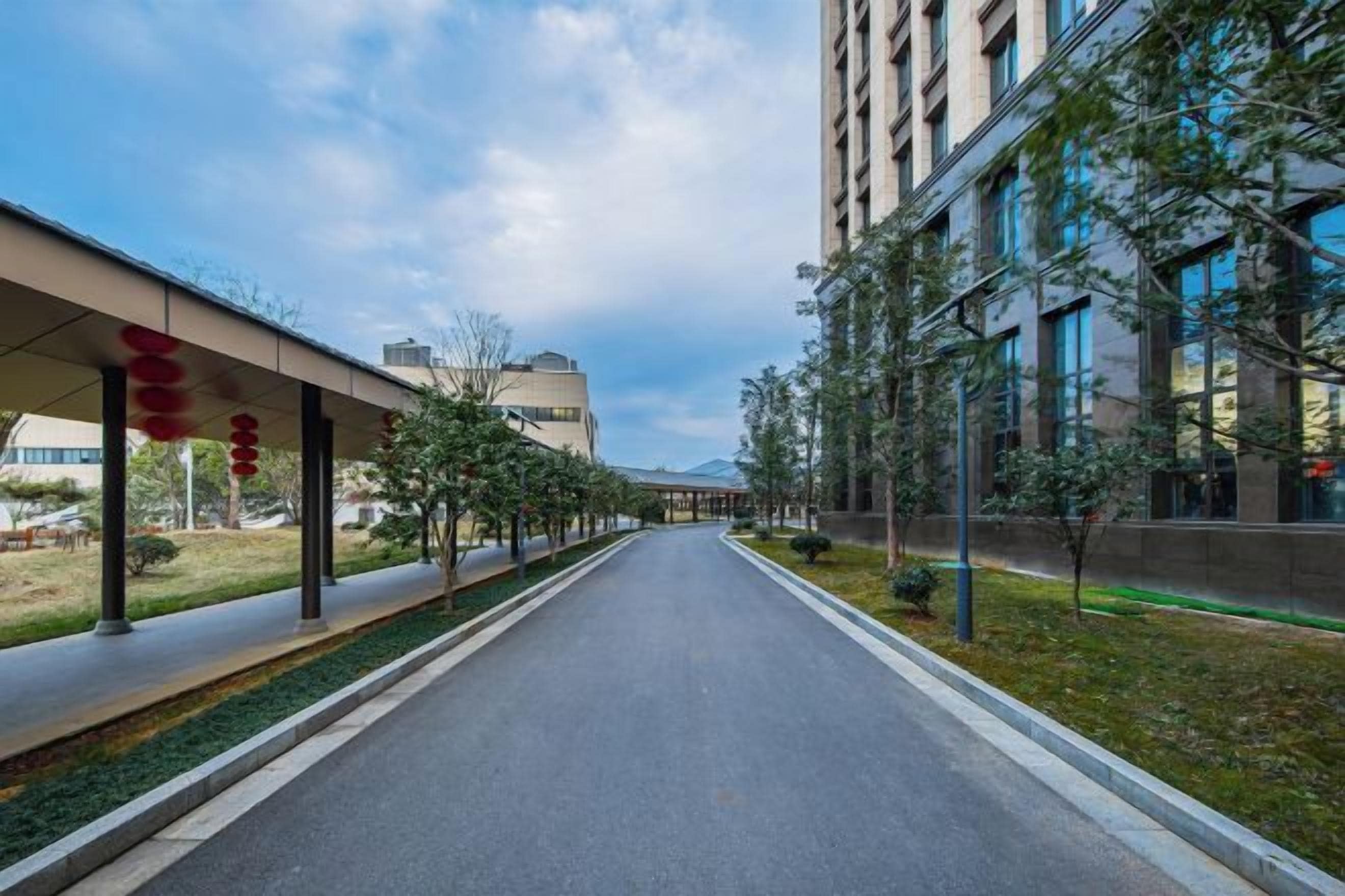 Un camino recto bordeado por edificios y árboles, que conduce a un edificio con techo plano y galería cubierta.