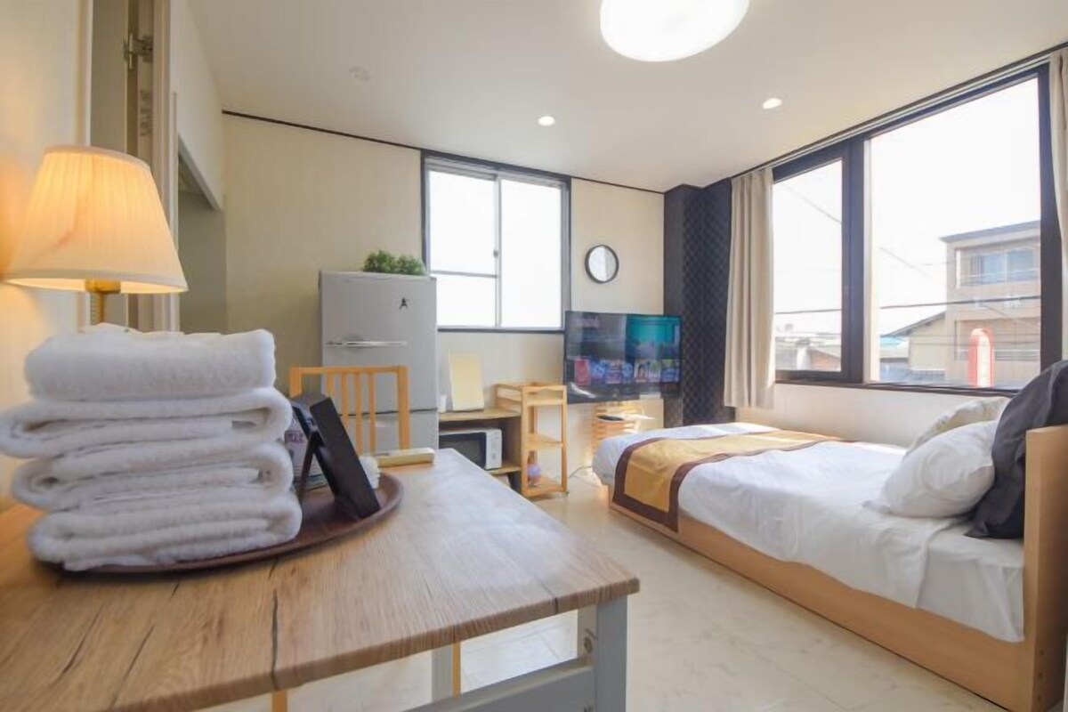 A hotel room with a bed, a desk with a lamp, a TV, and a window with a view of buildings.