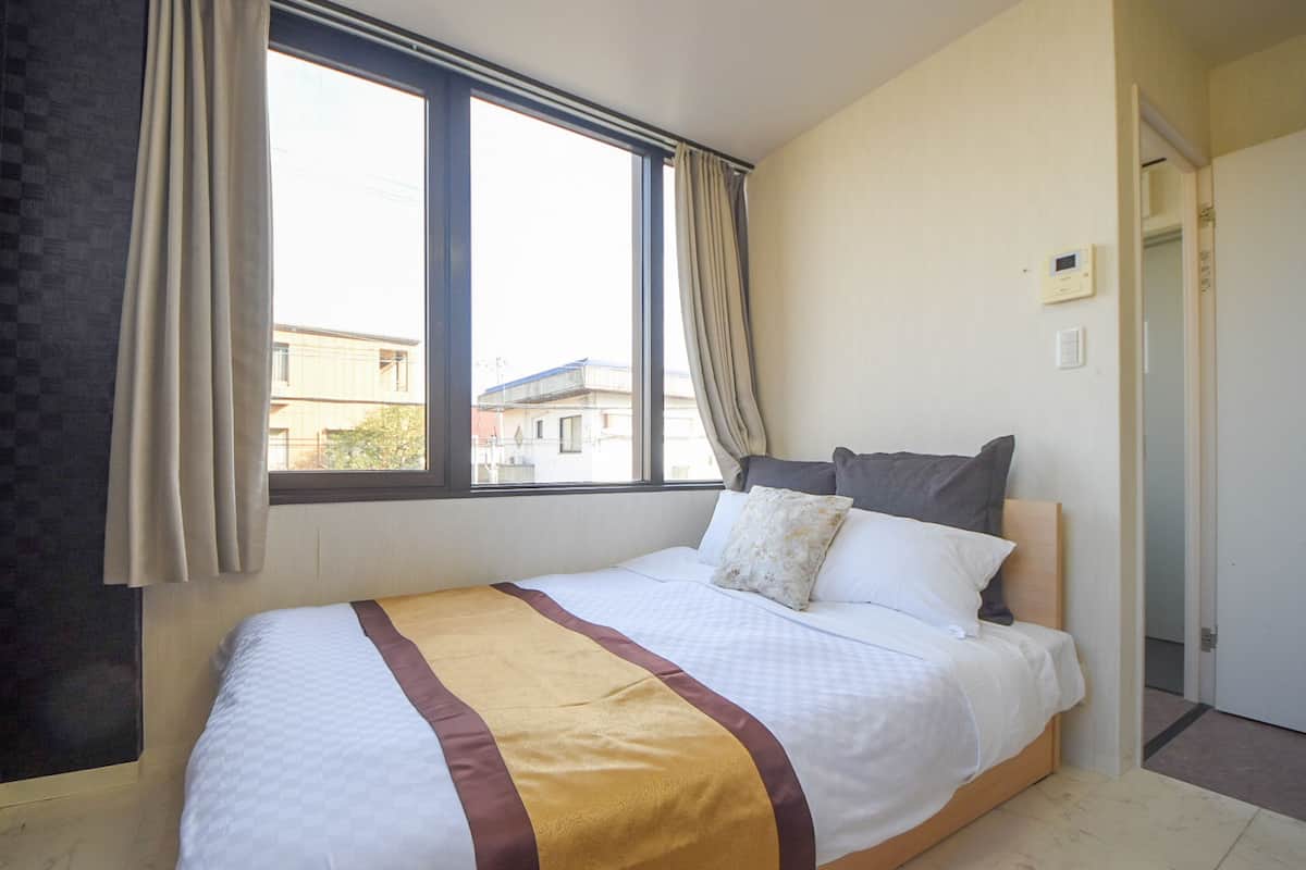 A hotel room with a large window, a bed with a white and brown bedspread, and a thermostat on the wall.