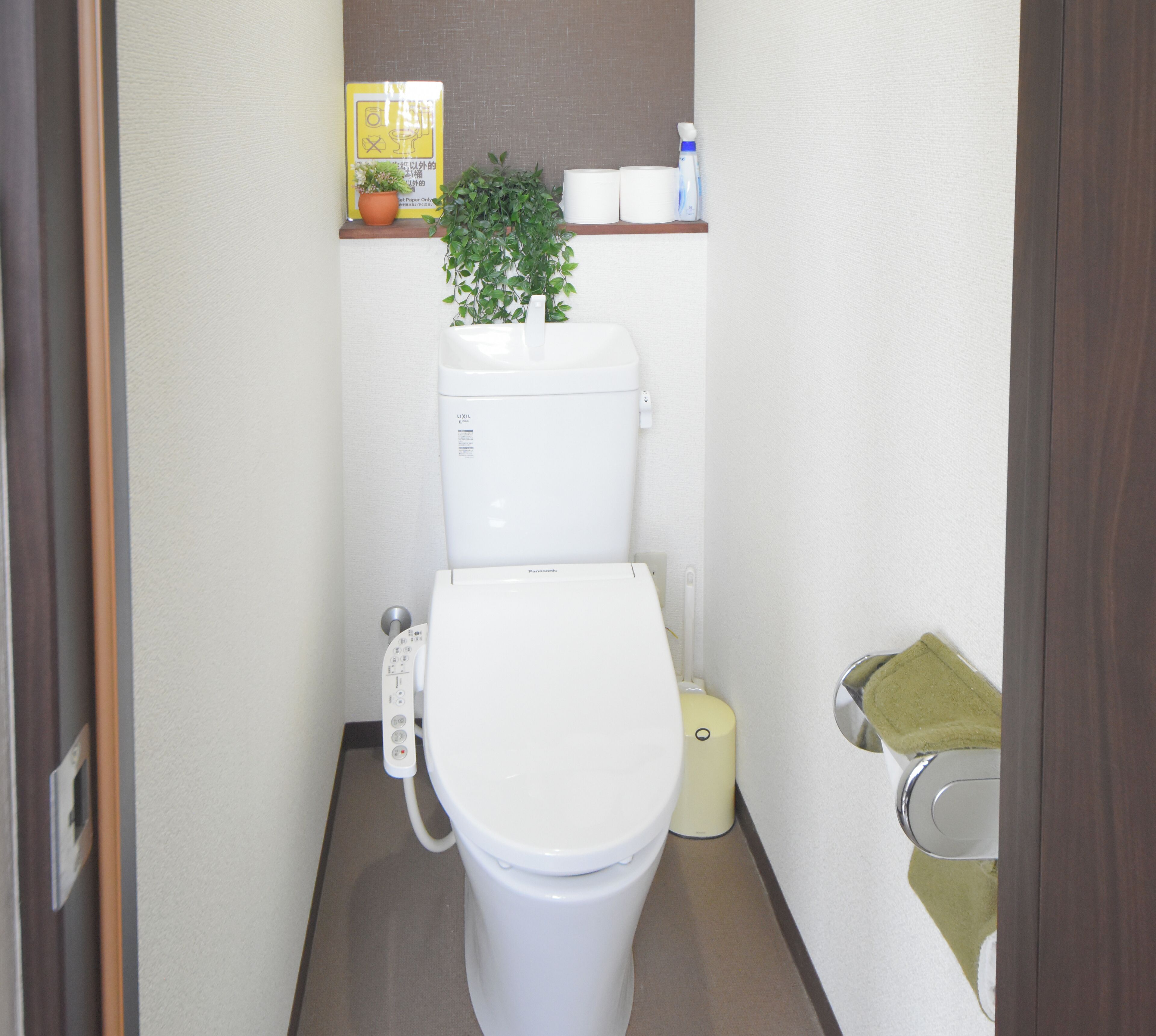 Ein kleines, sauberes Badezimmer mit Toilette, Mülleimer, Ablage für Toilettenartikel und einer Topfpflanze.