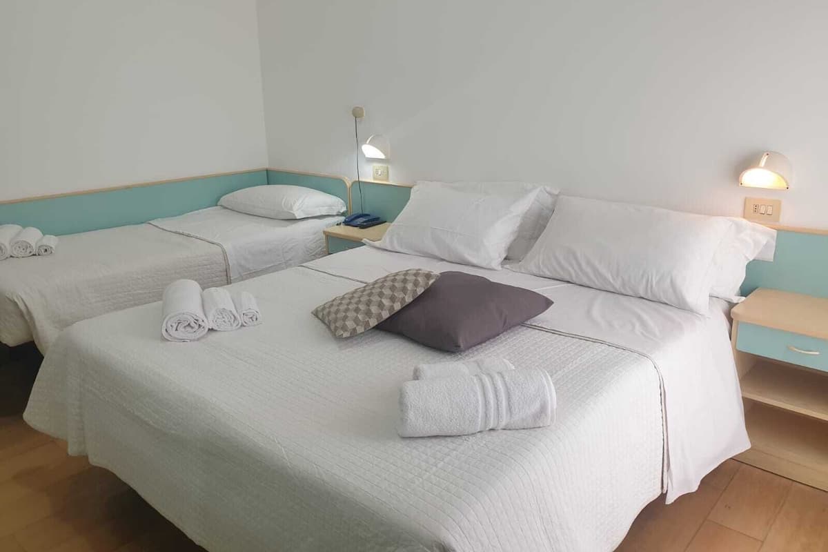 Ein Hotelzimmer mit zwei Einzelbetten, jeweils mit Kissen und zusammengerollten Handtüchern. An den Betten stehen Nachttische mit Lampen und ein kleines Regal.