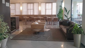 Un vestíbulo de hotel moderno con área de descanso, una recepción y ventanas grandes con cortinas.