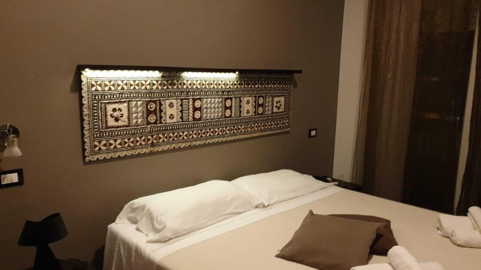 A hotel room with a bed, a patterned headboard, a nightstand with a lamp, and a window with curtains.