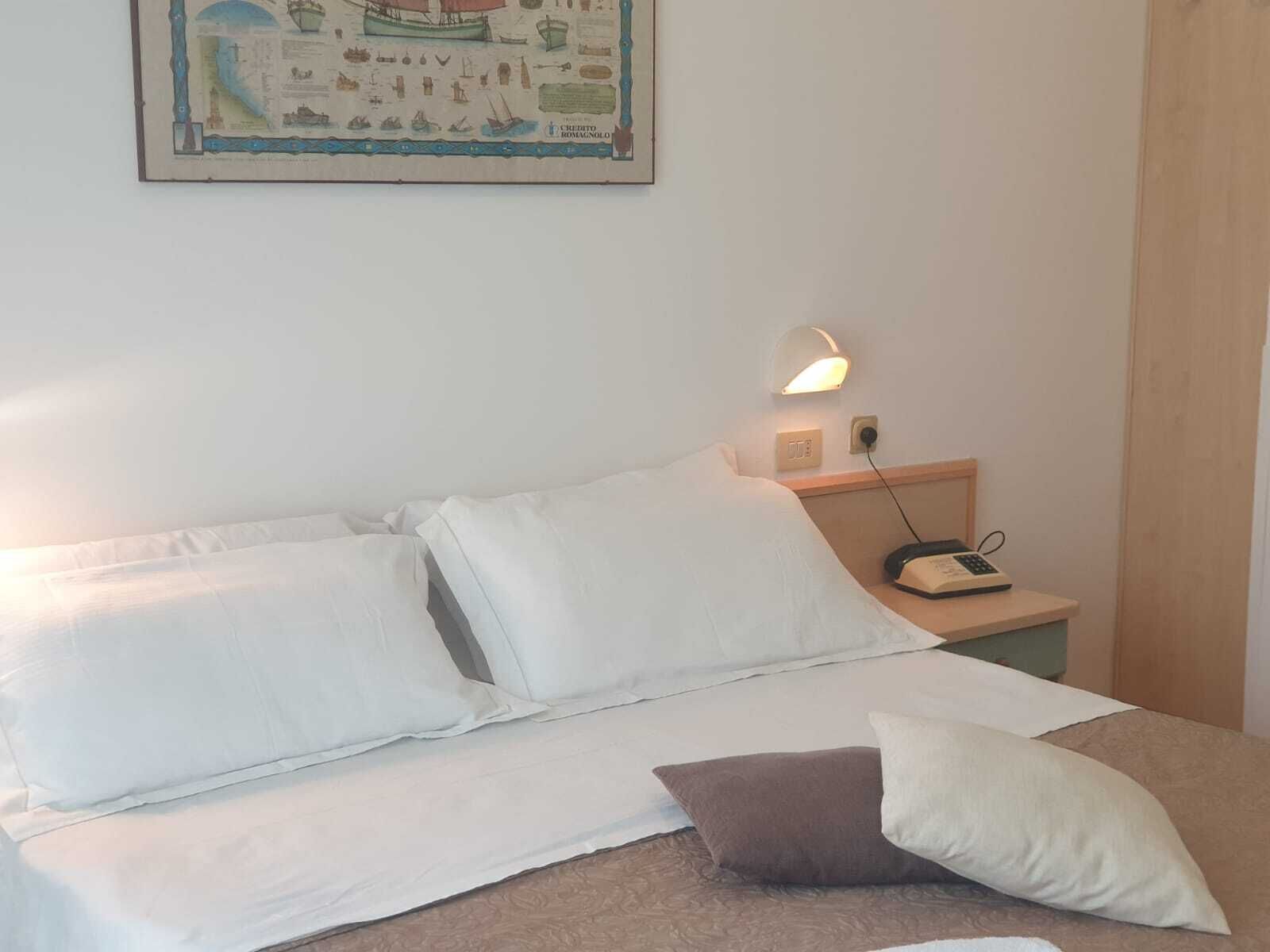 A hotel room with a bed, pillows, a nightstand with a lamp, and a wall-mounted painting.