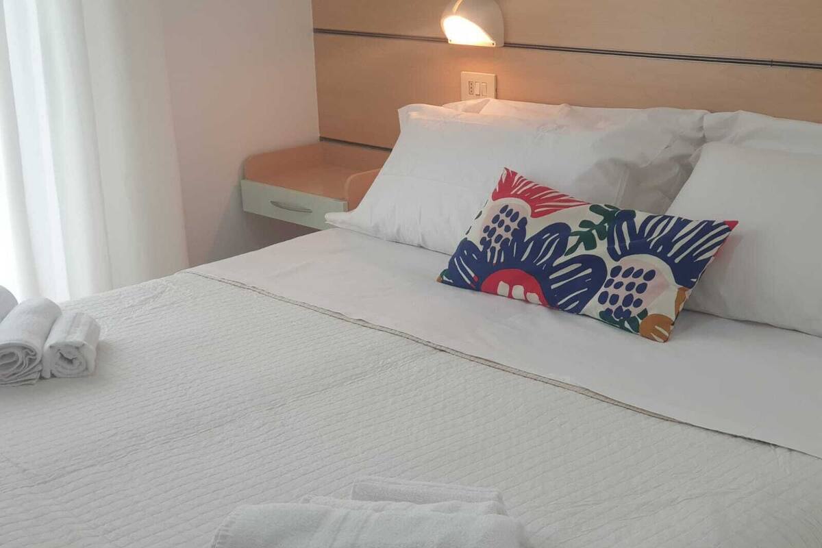 Ein ordentlich bezogenes Bett mit weißen Leinen, einem farbenfrohen Kissen und zusammengerollten Handtüchern daneben.