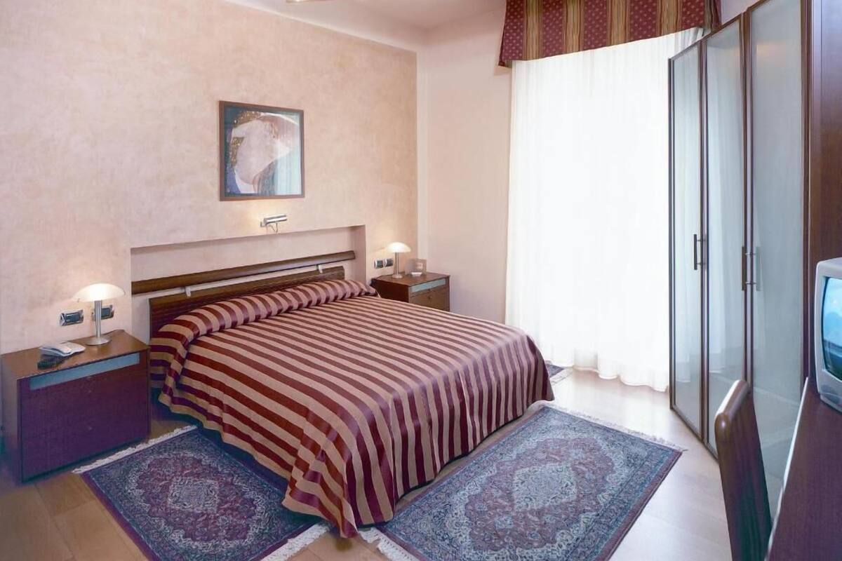 Ein Schlafzimmer mit Bett, Nachttischen, einem Schreibtischchen, einem großen Fenster mit Vorhängen und einer Deckenleuchte.