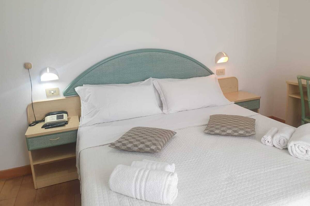 Ein ordentlich bezogenes Bett mit weißen Leinen, zwei Kissen und zusammengerollten Handtüchern. Ein Nachttisch mit Lampe, Telefon und einer kleinen Ablagefläche. Der Raum hat ein schlichtes, sauberes Design mit einem türkisblauen Kopfteil und hellen Wänden.