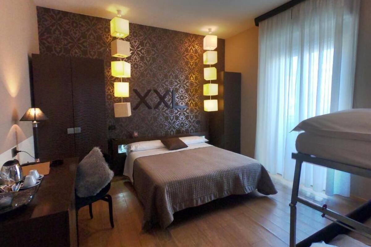 Ein Hotelzimmer mit einem großen Bett, Nachttischen, einem Schreibtisch und einem Stuhl. An den Wänden sind Lampen angebracht und es gibt ein Fenster mit Vorhängen.