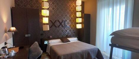 Quarto de hotel com uma cama grande, mesas de cabeceira, uma escrivaninha e uma cadeira. Há luminárias fixadas na parede e uma janela com cortinas.