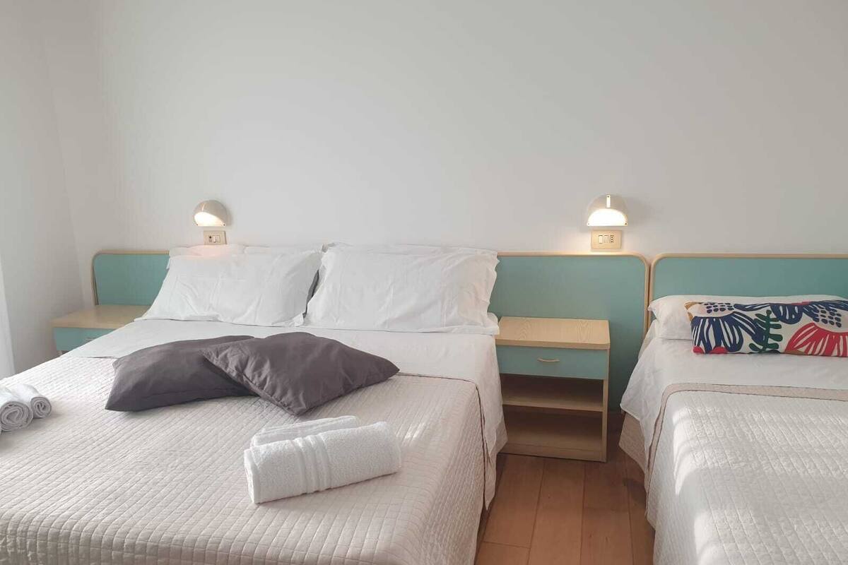 Ein Doppelzimmer mit zwei Betten, jeweils mit Kissen und zusammengelegtem Handtuch. An den Nachttischen stehen Lampen und kleine Schubladen.