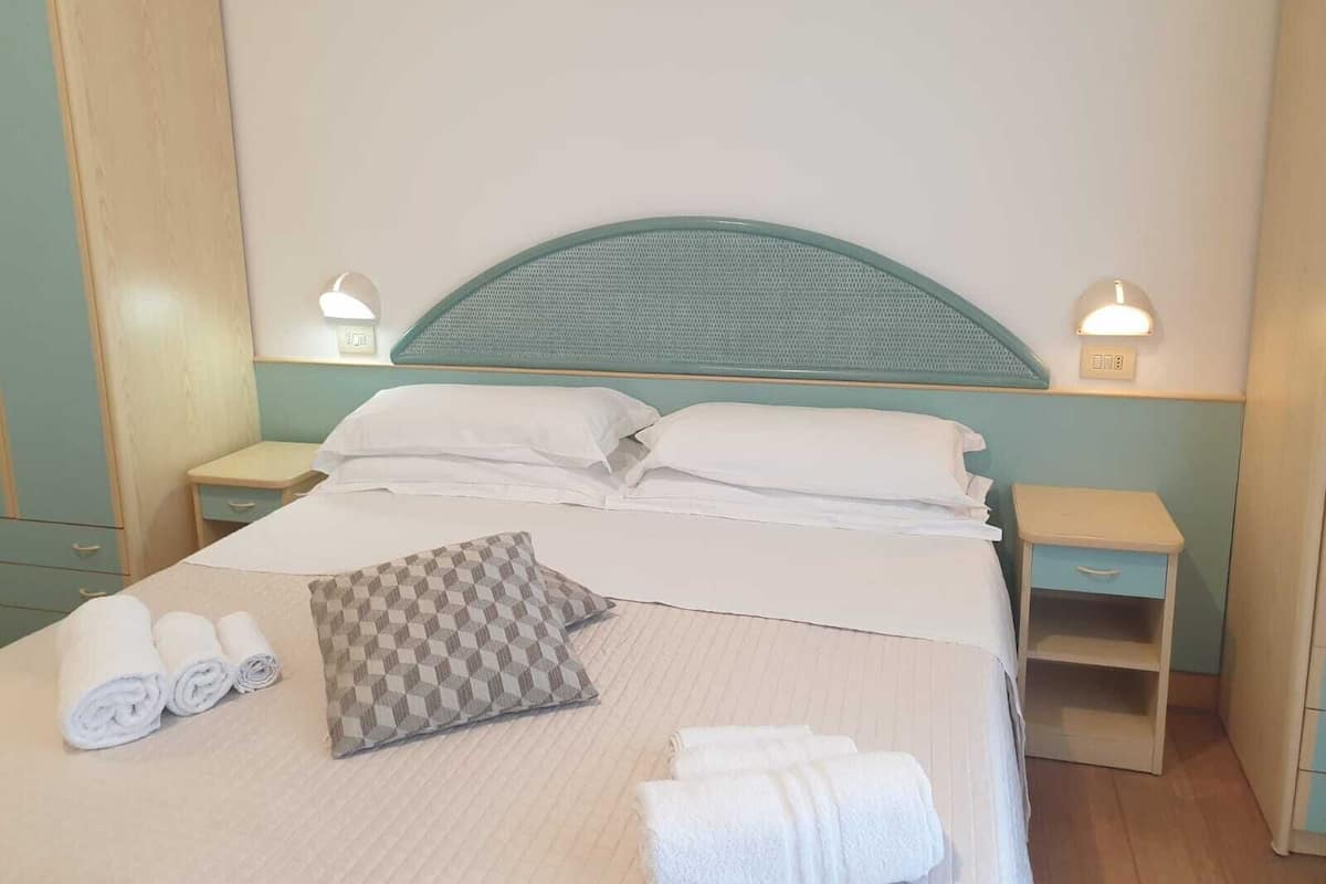 Ein ordentlich bezogenes Bett mit Kissen, darunter ein kariertes Kissen, und zusammengerollte Handtücher. Zwei Nachttische mit Lampen und eine kleine Ablagefläche sind ebenfalls vorhanden.