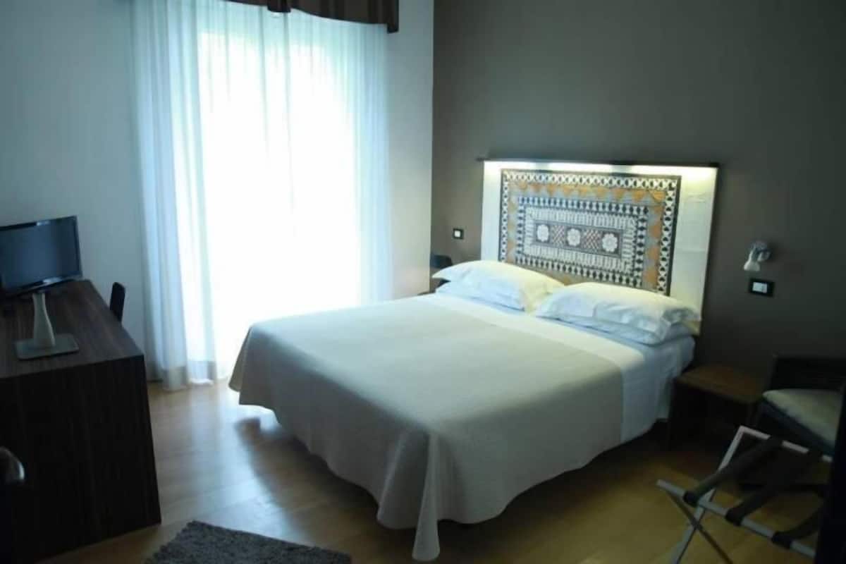 Ein Hotelzimmer mit einem Bett, einem Schreibtisch mit Stuhl, einem Fernseher und einem Fenster mit Vorhängen.