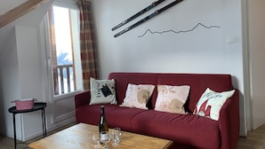 Un salon chaleureux avec un canapé rouge, une table basse en bois sur laquelle se trouvent une bouteille et des verres, une fenêtre avec des rideaux et des paires de skis accrochées au mur.