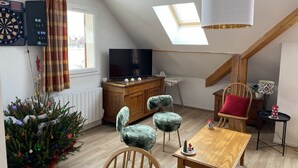 Une pièce chaleureuse de style loft, avec un plafond en pente, un téléviseur posé sur un meuble en bois, un petit sapin de Noël et une table à manger agrémentée d’une bougie rouge.