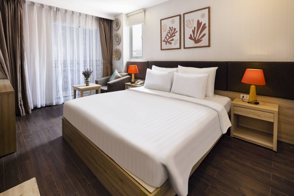A hotel room with a large bed, a bedside table with a lamp, a chair, a desk, and a window with curtains.