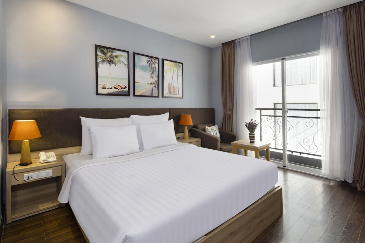 A hotel room with a large bed, a nightstand with a lamp, a chair, and a window with curtains.