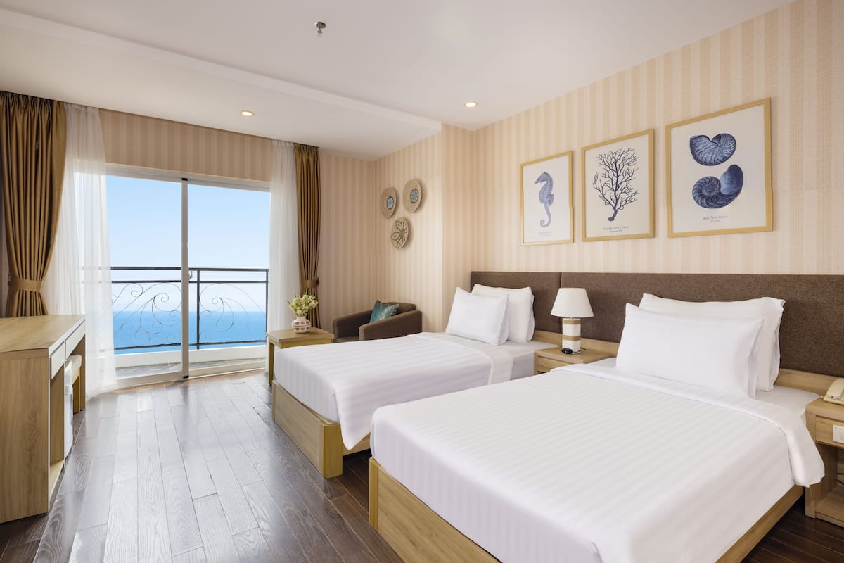 A hotel room with two beds, a desk, a chair, and a view of the sea.