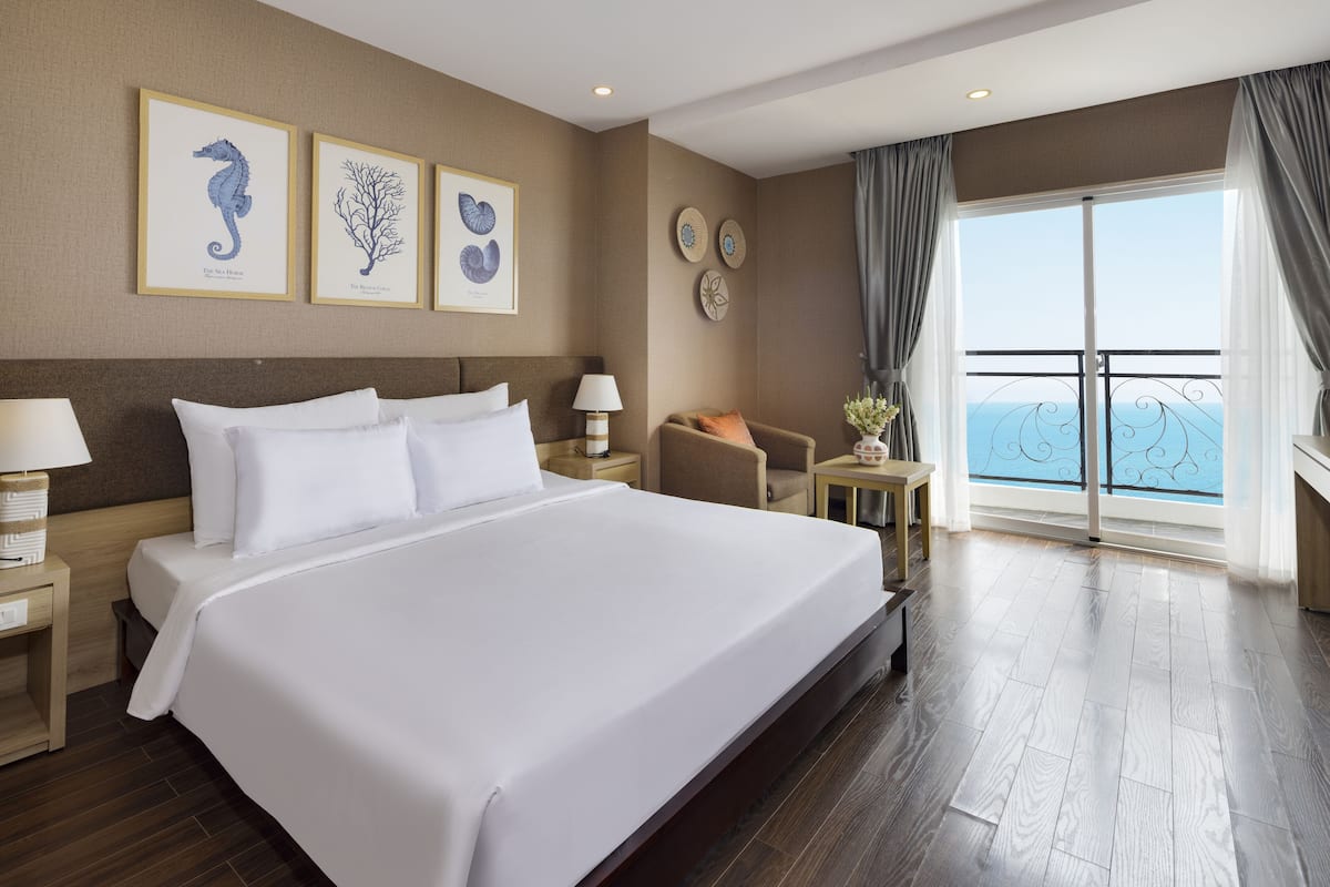 A spacious hotel room with a large bed, a nightstand, a chair, and a view of the sea through a balcony.
