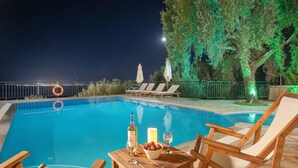 A poolside area with a table set for two, a bottle of wine, and a candle. There are lounge chairs and umbrellas around the pool. - Villa Kalithea in Nisaki (Corfu)