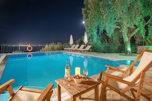 A poolside area with a table set for two, a bottle of wine, and a candle. There are lounge chairs and umbrellas around the pool. - Villa Kalithea in Nisaki (Corfu)
