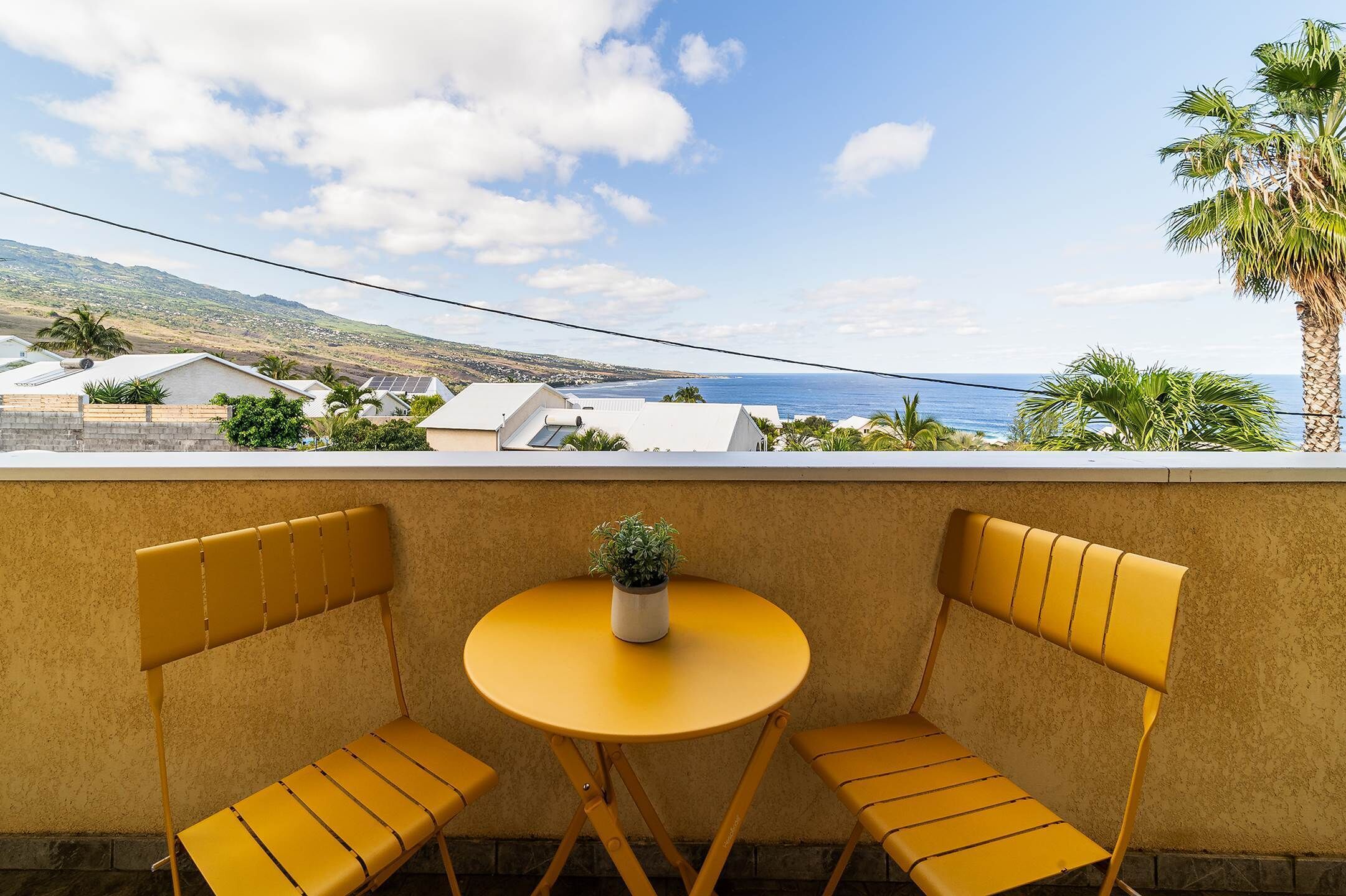 A yellow table and two yellow chairs with a small potted plant on the table, set against a wall with a view of the sea and distant hills.