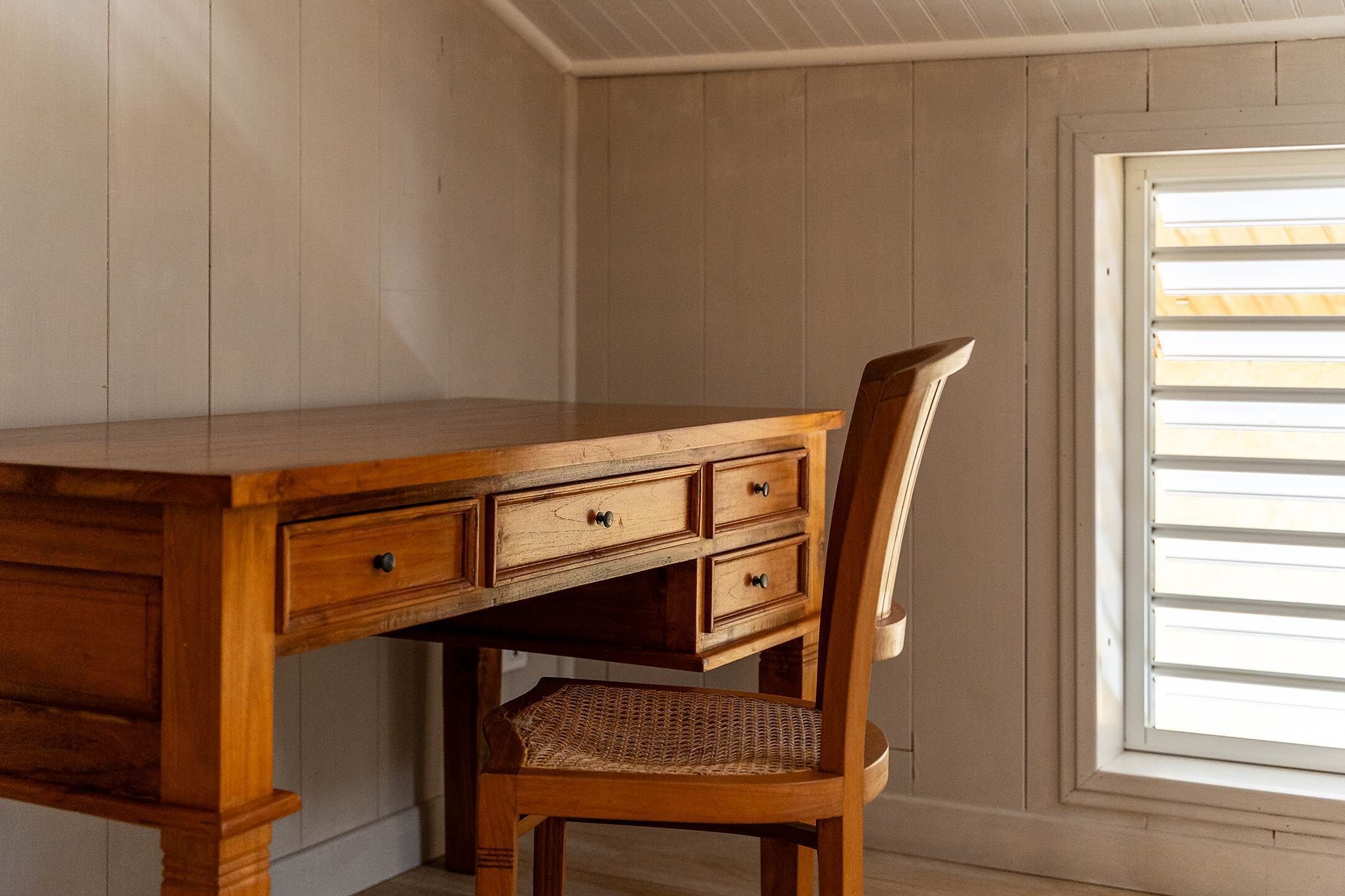 A wooden desk with drawers and a wicker chair in a room with wooden walls and a window with blinds.