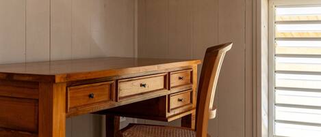 A wooden desk with drawers and a wicker chair in a room with wooden walls and a window with blinds.