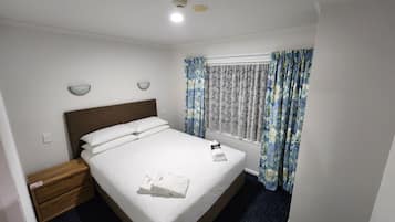 Una cama bien hecha con sábanas blancas, cabecera de madera y mesita de noche con lámpara.