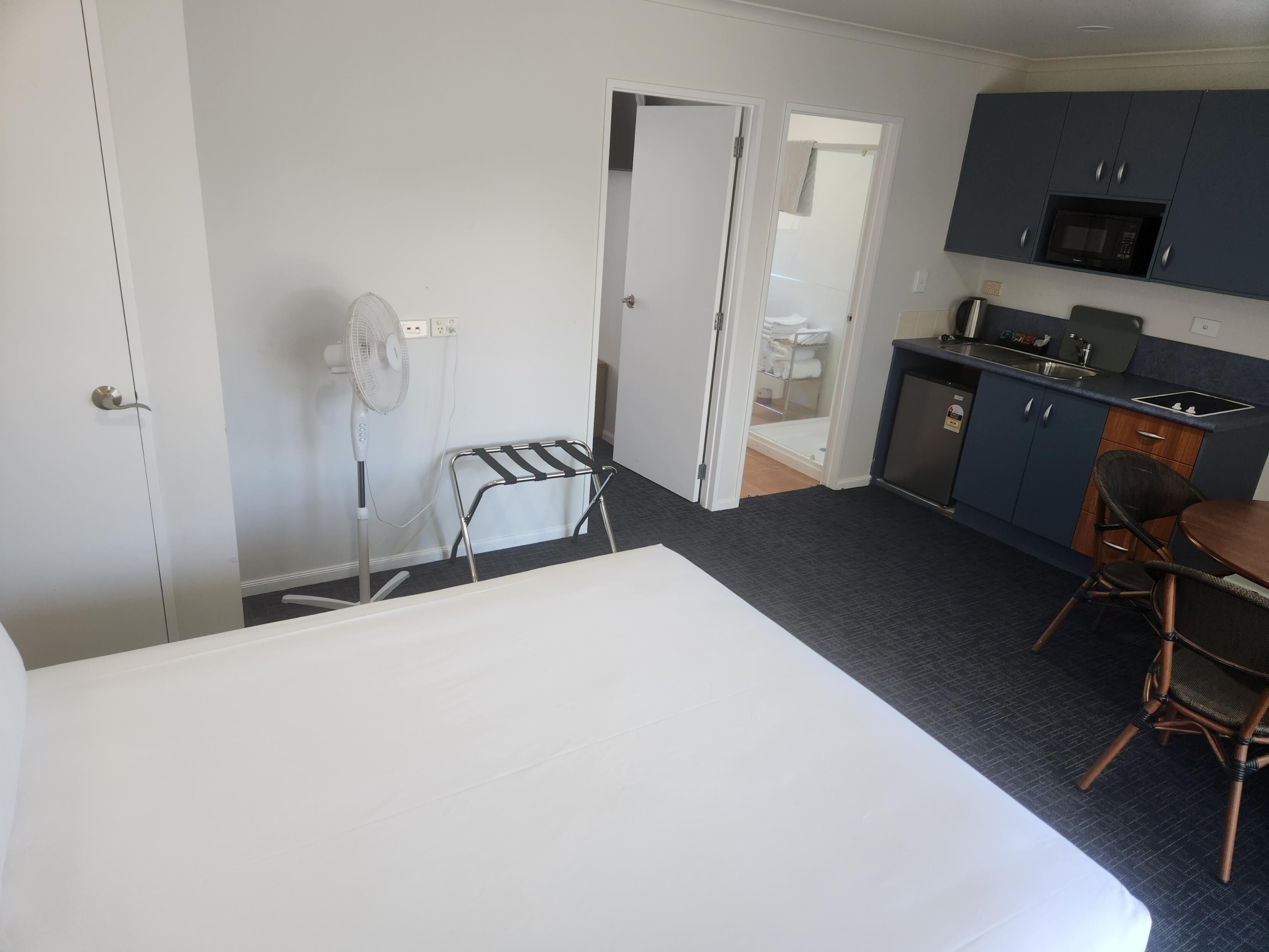 A hotel room with a bed, a small table, a chair, a fan, a microwave, and a kitchenette.
