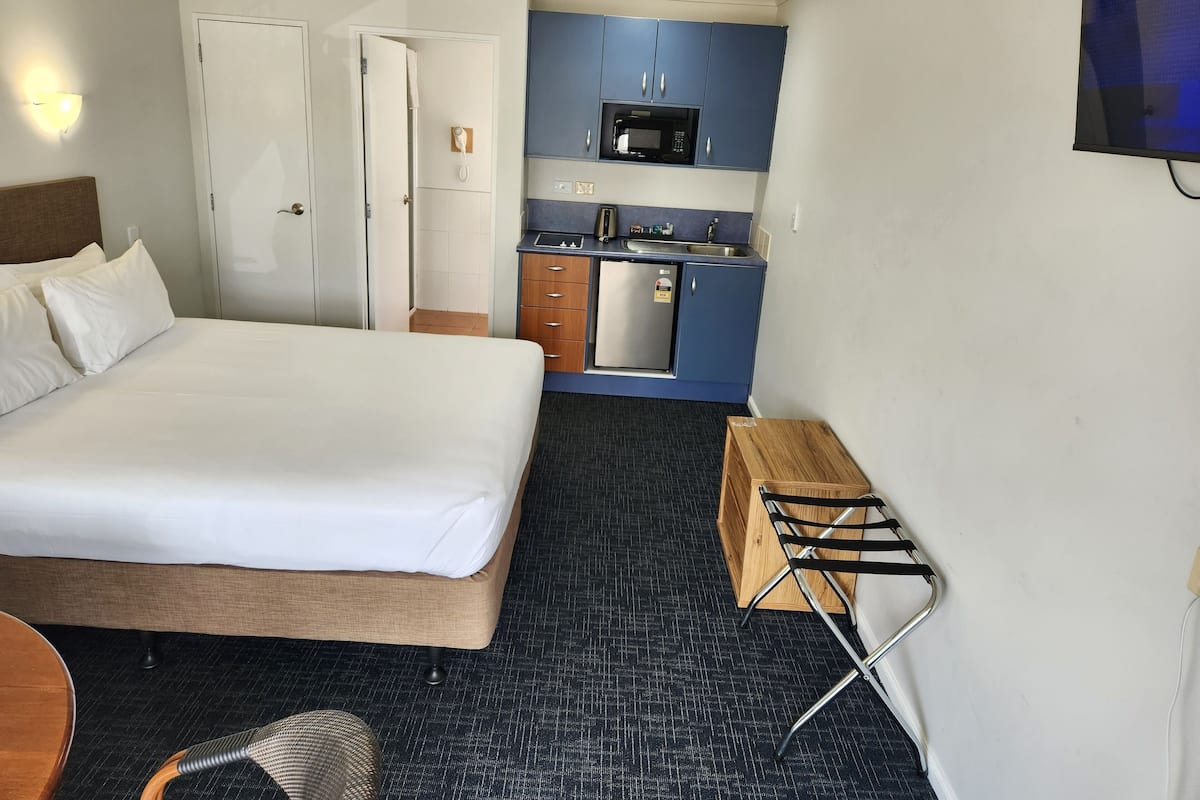 A compact hotel room with a bed, a kitchenette, a small wooden table, and a wall-mounted TV.