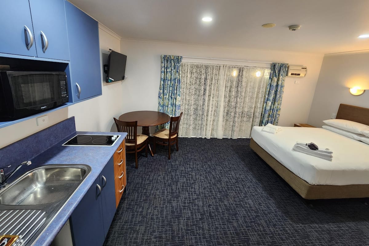 A compact hotel room with a kitchenette, a single bed, a small dining table, and a TV.