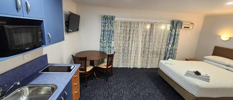 Habitación de hotel compacta con kitchenette, una cama individual, una pequeña mesa de comedor y un televisor.