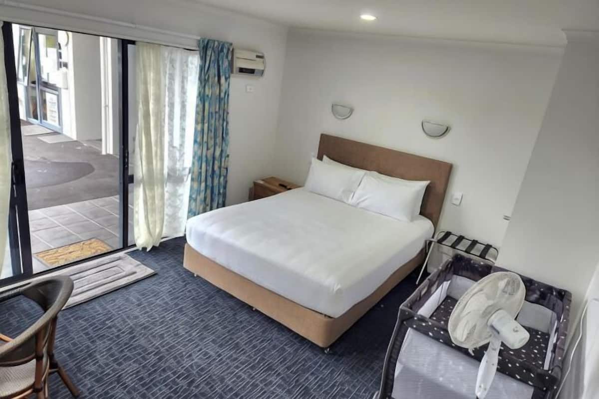 A hotel room with a large bed, a desk with a chair, a small table with a fan, and a sliding glass door leading to an outdoor area.