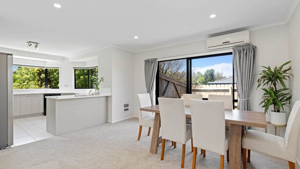 A modern kitchen with white cabinets, a central island, and a dining area with a wooden table and white chairs. - Botany Palms Motel (Auckland)