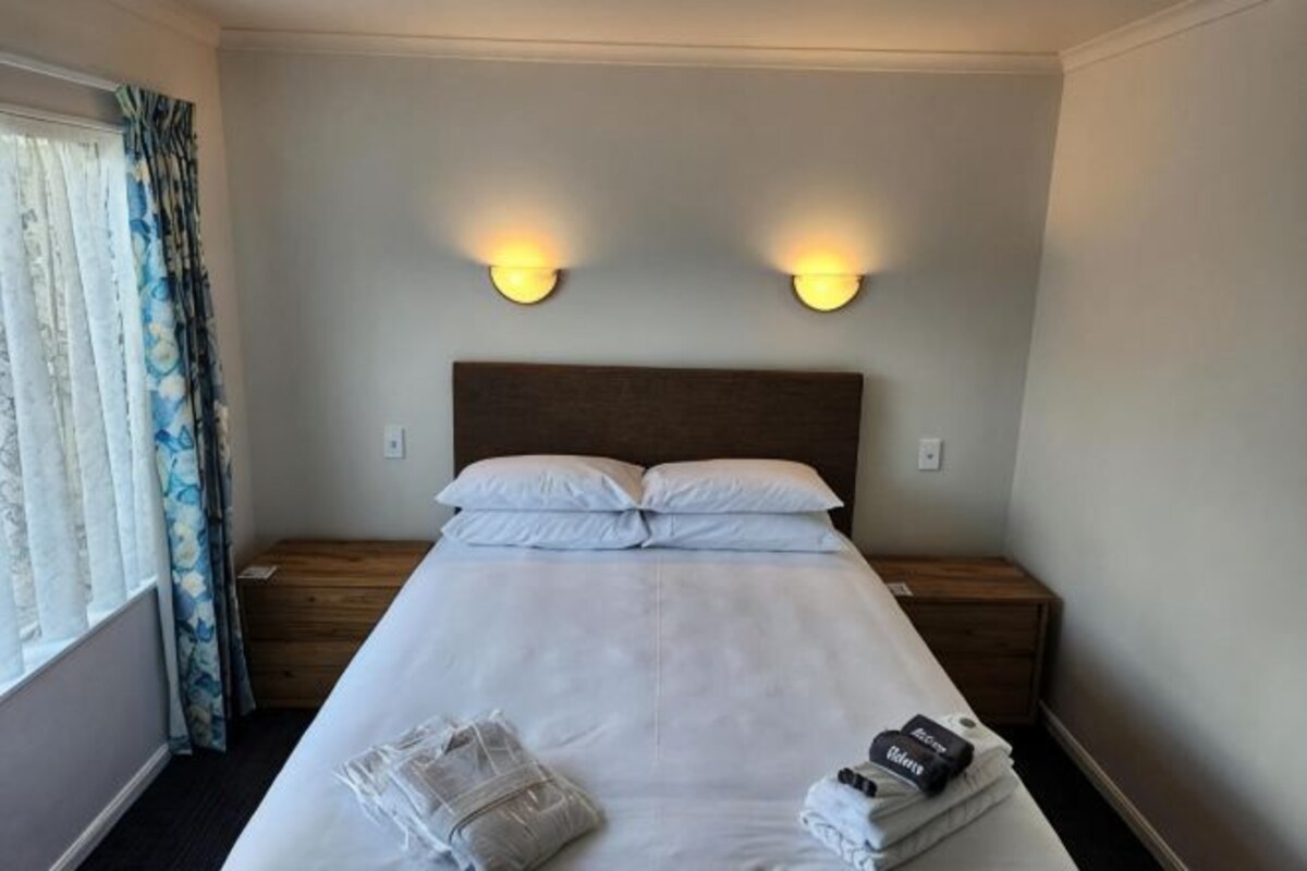 Una habitación de hotel con una cama, dos lámparas de noche, una ventana con cortinas y una mesita de noche con lámpara.