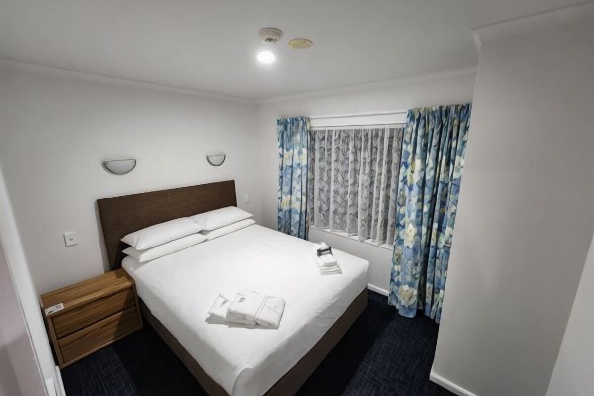 Una habitación de hotel con cama, mesita de noche, ventana con cortinas y lámpara fijada a la pared.