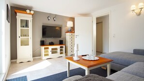 Un salon moderne avec un téléviseur à écran plat, un meuble blanc aux portes en verre, une table en bois sur laquelle est posée une bougie blanche, et des canapés gris recouverts de tissu.