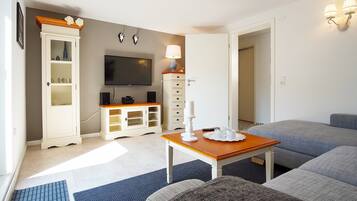 Ein modernes Wohnzimmer mit einem Flachbildfernseher, einem weißen Schrank mit Glastüren, einem Holztisch mit einer weißen Kerze und grauen Polstersofas.
