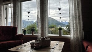 Ein Zimmer mit Blick auf Berge durch große Fenster, ein rotes Sofa, ein Holztisch mit Tassen und einem Korb, sowie ein Blumentopf.