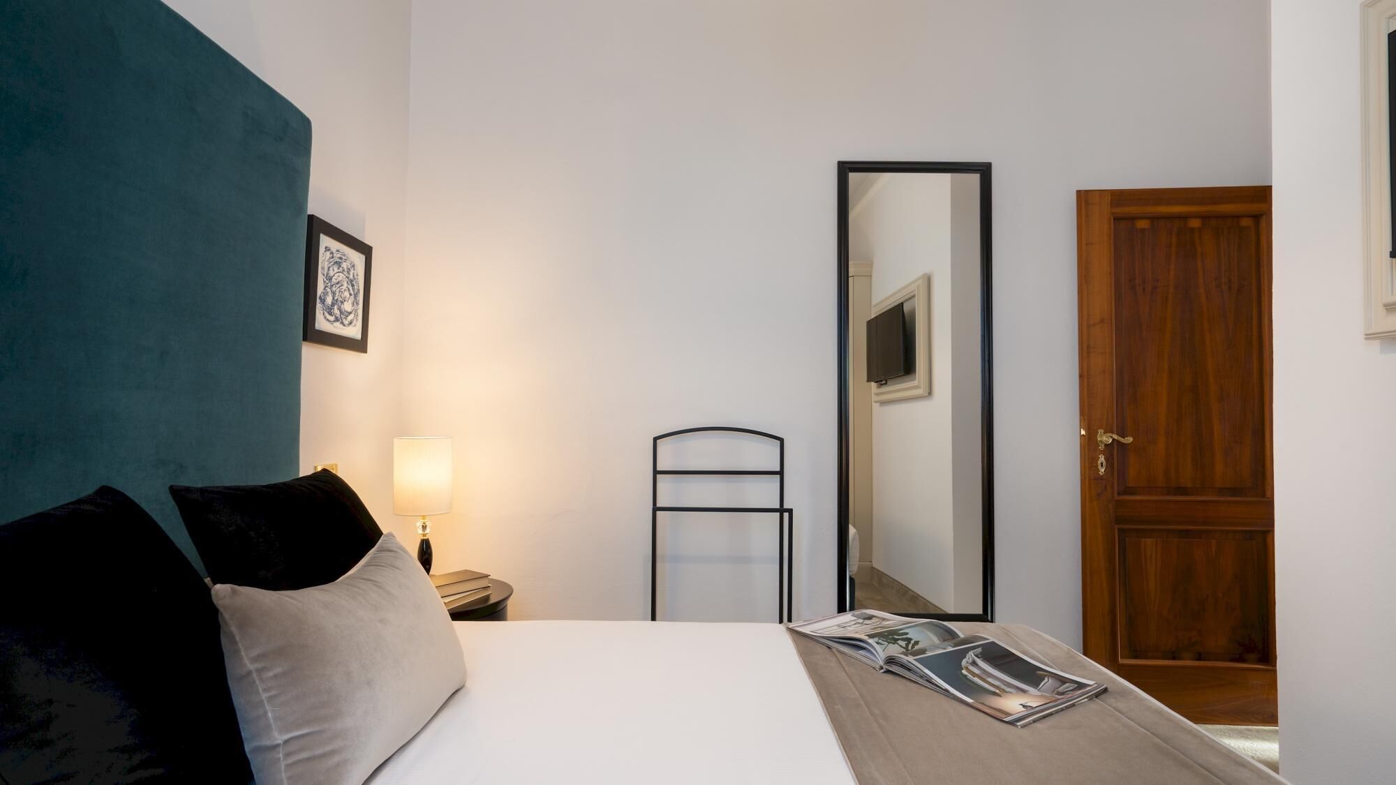 Un dormitorio con cama, una mesita de noche con lámpara, un espejo grande y una puerta de madera.